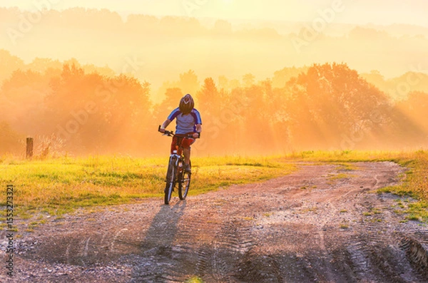 Fototapeta A person is riding a bike on a dirt road. The road is surrounded by trees. The sun is shining brightly, and the sky is orange