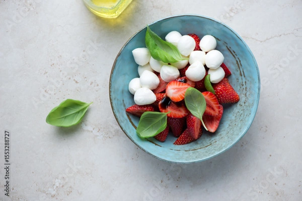 Fototapeta Strawberry caprese with mini mozzarella and green basil in a turquoise bowl, horizontal shot on a light-beige stone background, top view