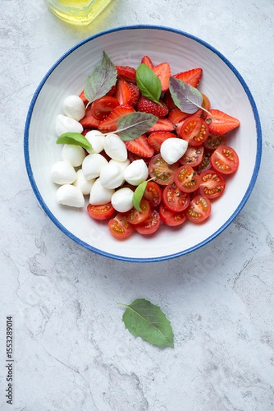 Fototapeta Plate of caprese salad with strawberry, cherry tomatoes, mozzarella cheese and fresh basil, vertical shot on a white stone background, above view
