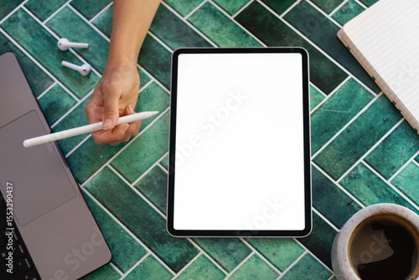 Obraz Top view of a woman’s hand holding a stylus above a digital tablet with a blank screen, surrounded by a notebook, coffee, and phone on green tiles. Perfect for creative work, digital drawing