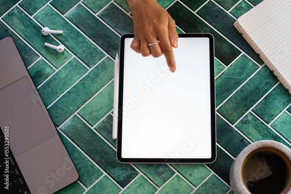 Obraz Flat lay of a woman's hand interacting with a digital tablet screen, surrounded by a notebook, stylus, smartphone, and coffee on a vibrant green tiled surface. Ideal for digital work concepts.