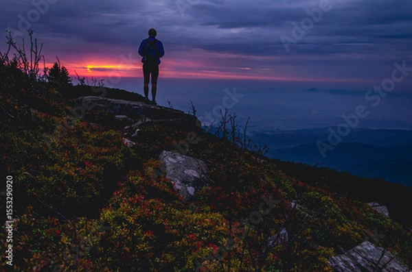 Fototapeta A man is standing on a hill with a backpack. The sky is orange and the sun is setting