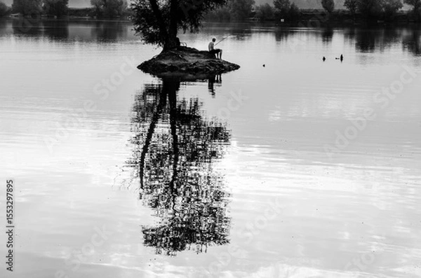 Fototapeta A man sits on a rock in front of a body of water. The water is calm and the reflection of the man and the rock can be seen in the water