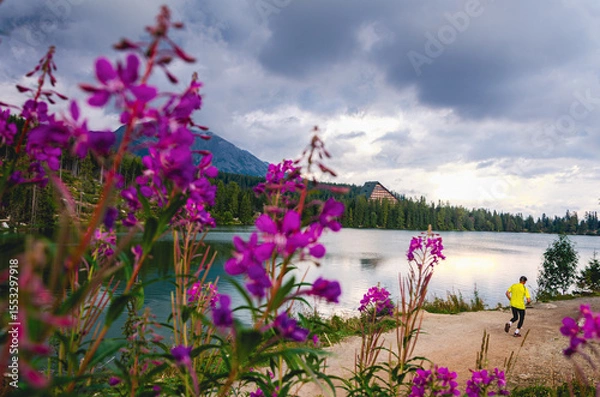 Fototapeta A man is running on a path near a lake. The lake is surrounded by purple flowers. The sky is cloudy and the man is wearing a yellow shirt
