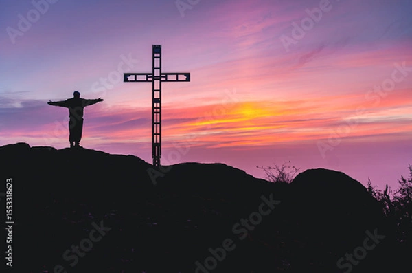 Fototapeta A man is standing on a hill with a cross in the background. The sky is a beautiful mix of pink and purple