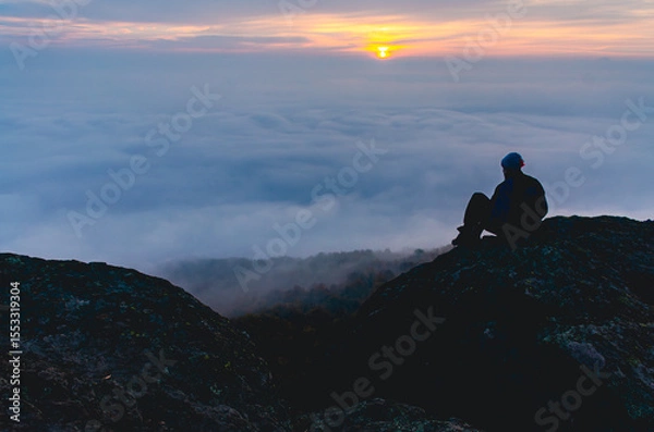 Fototapeta A man sits on a rock overlooking a foggy mountain. The sky is orange and the sun is setting