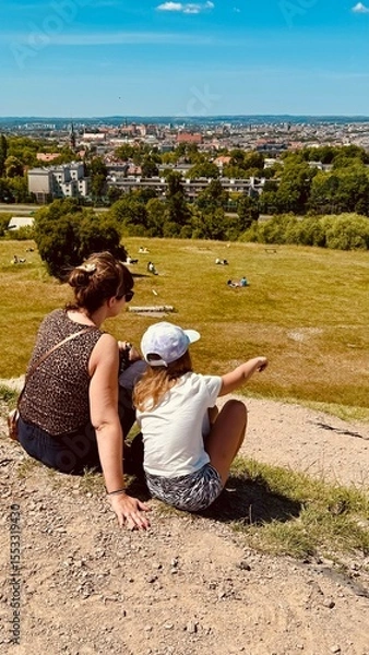 Fototapeta Mother and daughter sitting on hill with view on city of Cracow. Two people observe landscape among nature and park. Outdoors.