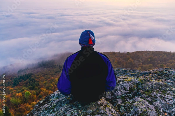 Fototapeta A man is sitting on a rock with a hat on. The sky is cloudy and the man is looking at the clouds