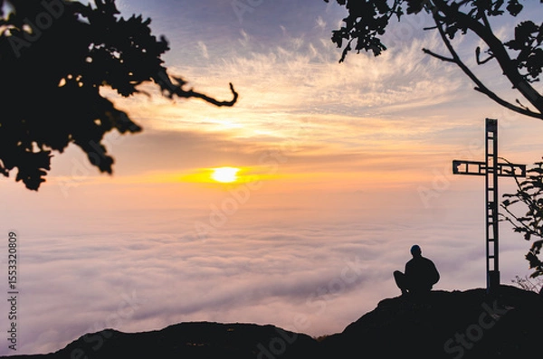 Fototapeta A man sits on a mountain top with a cross in the background. The sky is orange and the clouds are white