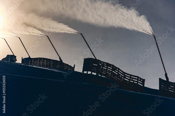 Fototapeta A group of snow guns are shooting water into the air, creating a misty atmosphere. The snow guns are positioned on a hill, possibly in a ski resort or a snow-covered area