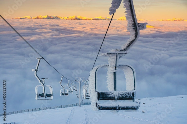 Fototapeta A ski lift with a chair covered in ice. The chair is located on the top of the lift, and it is surrounded by snow. The lift is located in a mountainous area, and the sky is cloudy