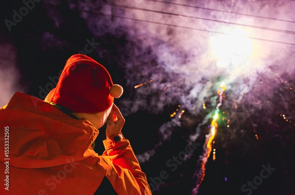 Fototapeta A person in a red hat is watching fireworks. The fireworks are in the sky and are colorful