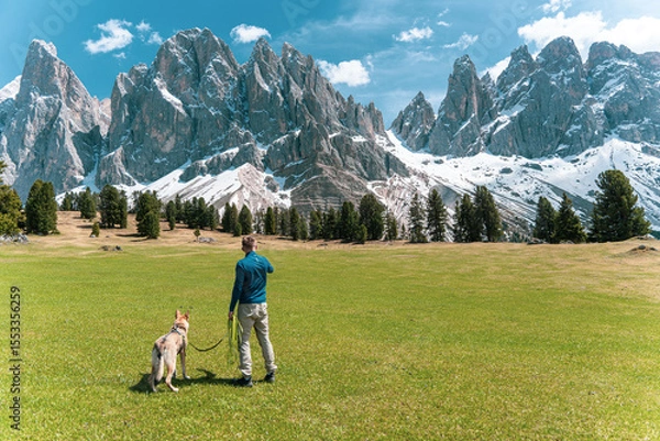 Fototapeta Man with dog standing on green meadow looking at snowy peaks in the Dolomites, Italy.