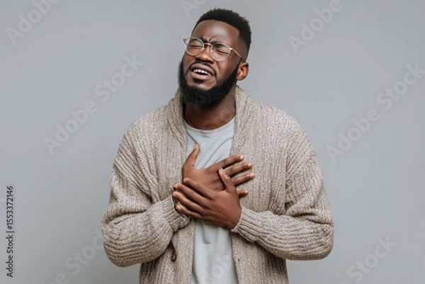 Fototapeta A man in a cardigan sweater appears to be experiencing a heart attack, clutching his chest in pain, isolated against a grey background