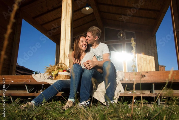 Fototapeta happy pretty couple in jeans and T-shirts is sitting and smiling on the step on the wooden house background on the sunset picnic, young family concept, free space
