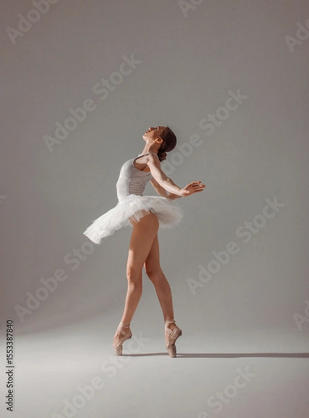 Fototapeta young slim dancer, fit girl ballerina stands in white ballet tutu like white swan in profile in pointe shoes on white wall background in studio light. classic ballet posing. ballet concept, free space