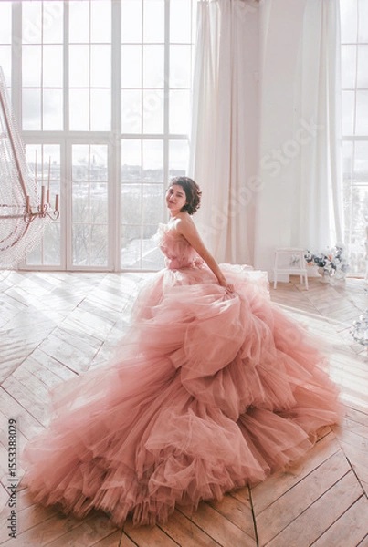 Fototapeta young girl with short black hair in pink princess dress with train is standing from the back and smiling cute near huge panoramic white window background at sunny day. fashion concept, free space