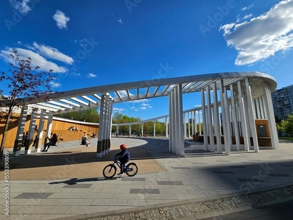 Fototapeta Young boy rides a bicycle in front of a large building construction gate in public park. There are several people in the background, some of them walking and others standing