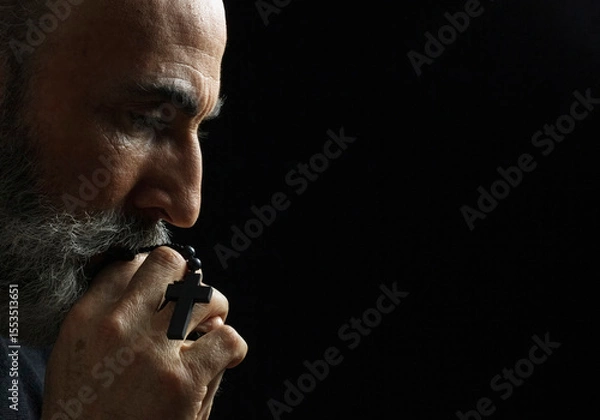Obraz Elderly man with gray beard holding rosary beads in prayer against dark background. Weathered hands and contemplative profile create powerful spiritual moment of faith and devotion.
