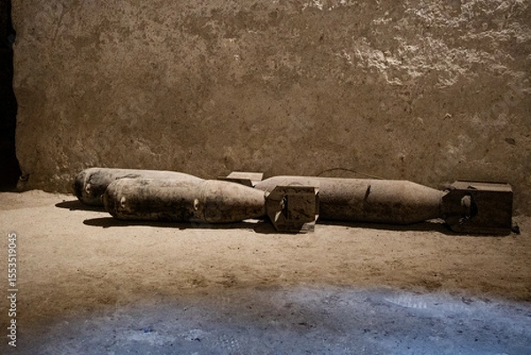 Obraz Weathered unexploded WWII bombs lying on dusty ground against textured concrete wall. Historical military ordnance creates powerful wartime atmosphere.
