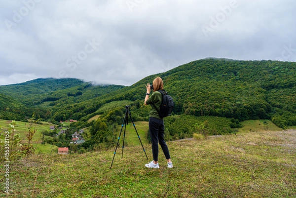Obraz Woman photographer with a tripod capturing a stunning mountain landscape and a village below. Female traveler taking photos of a scenic mountain valley on a cloudy day in the Carpathians