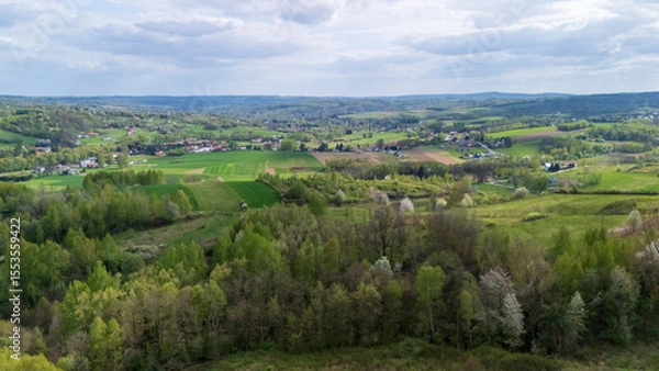 Obraz Aerial view of lush green fields and forests under a partly cloudy sky, showcasing the natural beauty of the countryside in spring, inviting outdoor exploration.