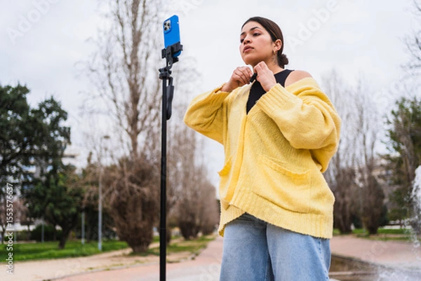 Fototapeta Latin influencer adjusting cardigan while filming content for social media with smartphone on tripod in a park