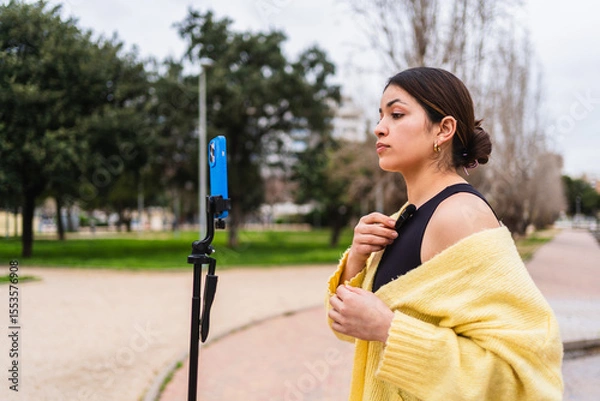 Fototapeta Young woman attaching her lavalier microphone while preparing to record a video with her smartphone on a tripod in a park