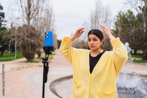 Fototapeta Young woman adjusting her hair while recording a video blog in a park, using a smartphone on a tripod with a lavalier microphone