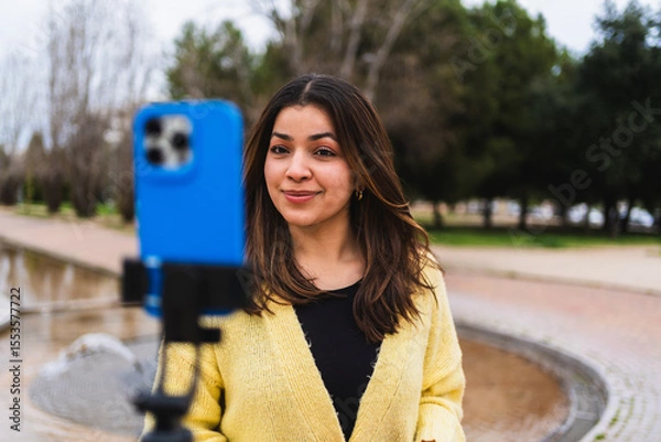 Fototapeta Young woman vlogger creating video content for social media using a smartphone and tripod in an urban park setting