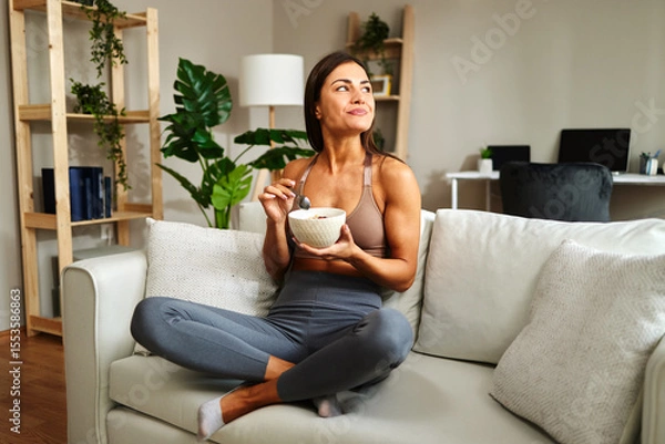 Obraz Young sportswoman eating healthy breakfast at home while sitting on sofa