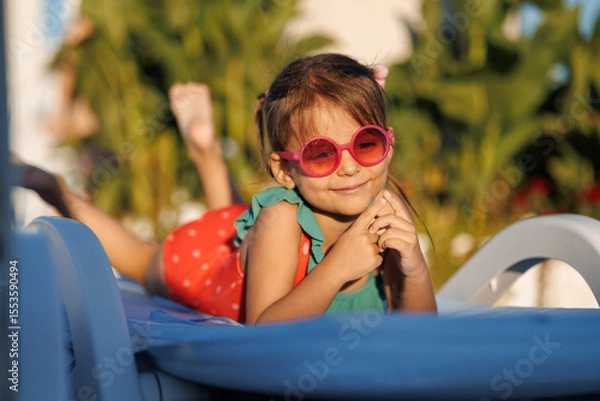 Fototapeta Funny little girl relaxing on summer vacation. The child has fun on the beach. Cute baby in a colorful swimsuit and sunglasses is resting.