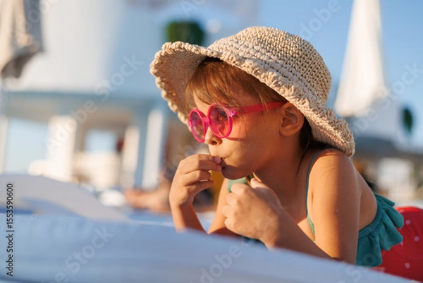 Fototapeta Funny little girl relaxing on summer vacation. The child has fun on the beach. Cute baby in a colorful swimsuit and sunglasses is resting.