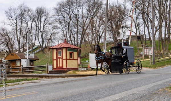 Fototapeta An Amish horse-drawn carriage is seen crossing a rural road adjacent to traditional buildings. The scene captures the charm of early spring with bare trees and a peaceful countryside atmosphere.