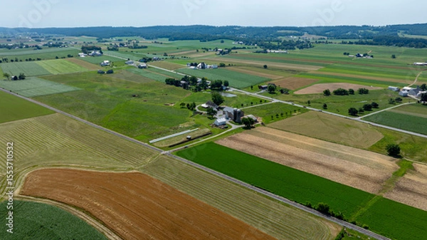Fototapeta A vast, open landscape showcases fields of varying colors and textures, with a few farmhouses scattered throughout. The clear sky allows for bright sunlight illuminating the fields.