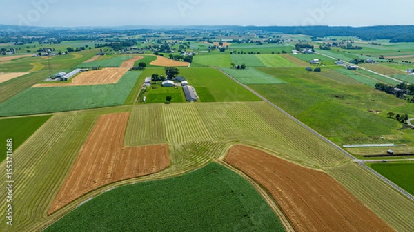 Fototapeta This expansive rural landscape reveals a patchwork of fields in various stages of crop growth. Rolling hills surround the area, with farm buildings dotting the horizon under a clear sky.