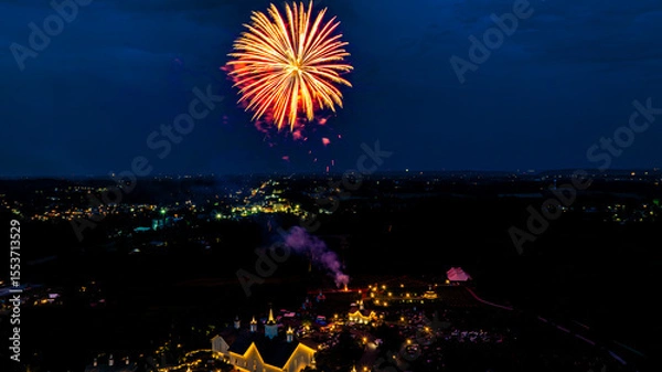 Fototapeta Colorful fireworks explode against a dark sky, illuminating a lively gathering below. The event takes place in a countryside setting, showcasing illuminated buildings and a festive atmosphere.