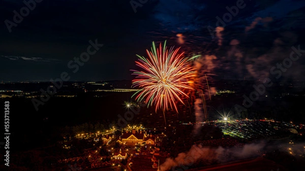 Fototapeta Colorful fireworks burst in the night sky over a lively event, with lights sparkling from the structures below. The atmosphere is filled with celebration as people enjoy the festivities.