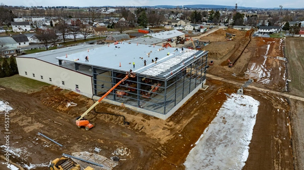 Fototapeta Workers are busy constructing a large industrial building in a suburban area. Heavy machinery is in use, and the ground is partly muddy with patches of snow present, indicating early spring weather.