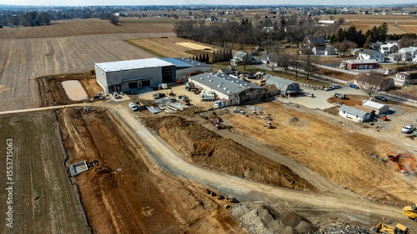 Fototapeta In a rural setting, work continues on a construction site featuring new buildings and road infrastructure. Equipment, vehicles, and materials are visible, showcasing active construction activities.