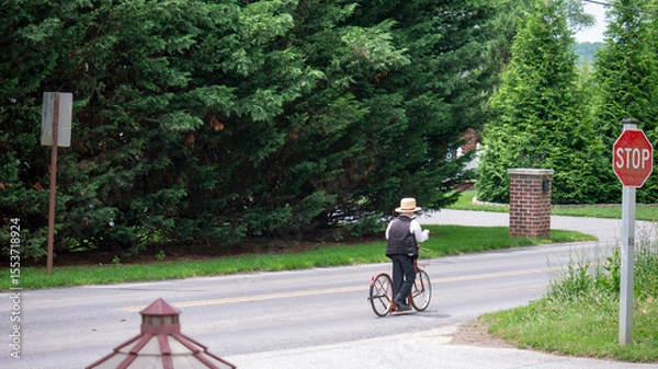 Fototapeta A Young Amish man dressed in traditional attire rides a vintage bicycle along a quiet rural road. Tall green trees line the roadside, while a stop sign signals caution nearby. Soft sunlight bathes the