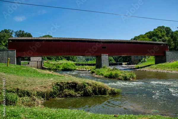 Fototapeta A historic covered bridge crosses over a calm river, surrounded by lush greenery and trees under a clear blue sky. The peaceful setting offers a glimpse into nature and architecture.