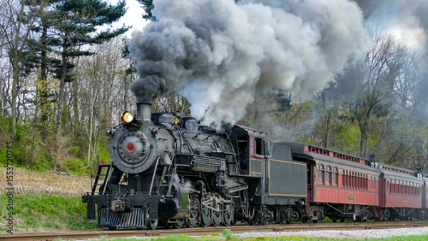 Fototapeta A historic steam locomotive travels along a scenic railway, releasing clouds of smoke into the air. The train is surrounded by lush greenery and trees, showcasing a vibrant spring landscape.