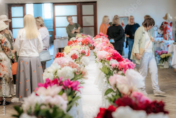 Fototapeta Rows of colorful peonies are displayed on long tables at a flower exhibition, with people in the background admiring the floral arrangements.