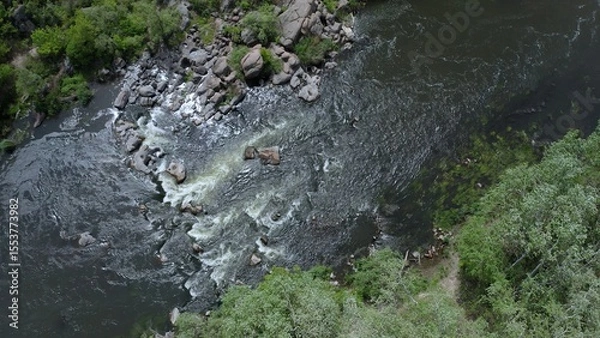Fototapeta Aerial view of rocks across a section of a flowing stream. Trees and large stone boulders around. Gusts of wind swaying tree branches.