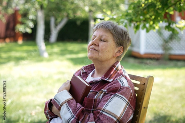 Fototapeta Senior woman in a checkered jacket enjoying a peaceful sunny day with closed eyes and holding a book to her chest, concept of relaxation and mindfulness in old age