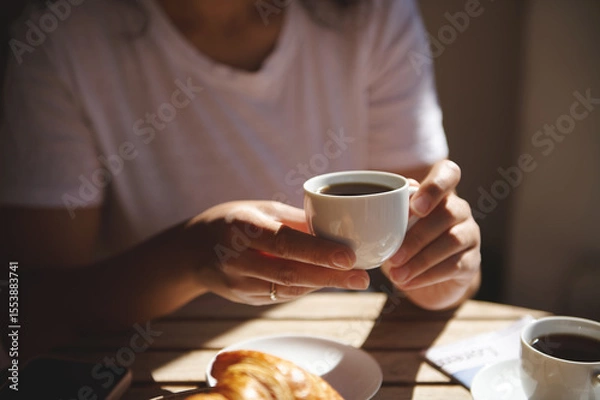 Fototapeta Close-up of hands holding a cup of coffee with a croissant on the table. A cozy moment enjoying breakfast in a bright and warm setting.