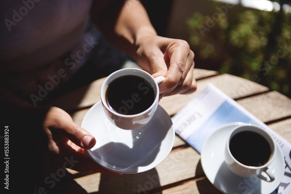 Fototapeta A person savoring a cup of coffee outdoors at a sunlit table, accompanied by a magazine and another cup. The peaceful ambiance underscores leisurely mornings and outdoor relaxation.