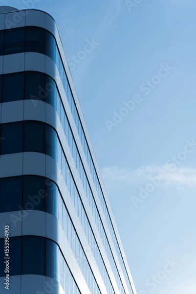 Fototapeta Contemporary office building with curved glass and metal facade, photographed against a clear blue sky. The image highlights modern architecture and urban business environments.