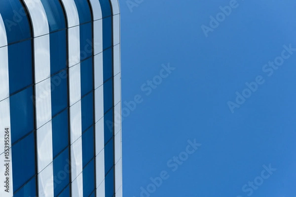 Fototapeta A contemporary office building with reflective glass windows is shown on the left, contrasted by a clear blue sky. The image highlights urban architecture and modern design for business or real estate